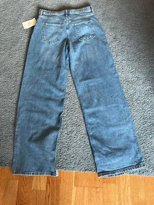 Blå raka jeans 170 - Säljer ett par klassiska blå jeans i rak modell med hög midja och två bakfickor. Jeansen har en avslappnad passform och är tillverkade i ett slitstarkt denimtyg. Perfekta för en trendig och bekväm look.