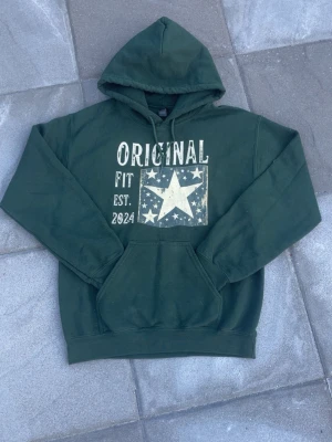 Original hoodie mörkgrön  - Tjoooo! Säljer en svin snygg Mörkgrön hoodie från Original Fit med stor vit stjärna och texten 'ORIGINAL FIT EST. 2024' på bröstet. Klassisk känguruficka framtill och huva. Tillverkad i mjukt bomullsmaterial, perfekt för en avslappnad laidback/grisch outfit🕺storlek S skick 9,5/10! Sjukt snygg🤩hör av dig vid mer frågor eller funderingar! Mvh Ga😁💫
