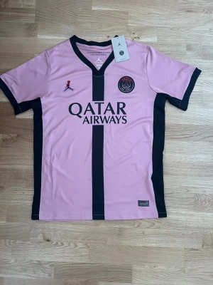 PSG x Jordan rosa matchtröja - Snygg rosa PSG matchtröja i samarbete med Jordan. Tröjan har svarta detaljer, V-ringning och klubbmärke på bröstet. Tillverkad i lätt och ventilerande funktionsmaterial, perfekt för fotboll eller träning. Qatar Airways-logga på framsidan.