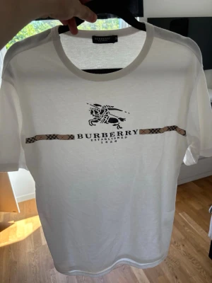 Vit Burberry t-shirt med logga - Snygg vit t-shirt från Burberry med klassisk logga och riddare framtill samt ett beige-rutigt band över bröstet. Tillverkad i 100% bomull och har en normal passform med korta ärmar. Perfekt för dig som gillar stilrena designer med exklusiv känsla.