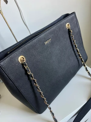 Svart handväska från DKNY med kedja - Snygg svart handväska från DKNY i strukturerat läder med guldfärgade detaljer och kedjeaxelband. Väskan har dragkedja upptill och en stilren, fyrkantig form. Perfekt för dig som gillar klassisk och elegant design med en modern twist.