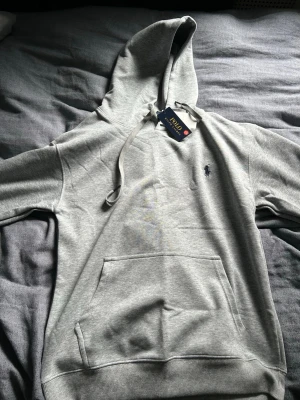 Grå hoodie från Polo Ralph Lauren - Snygg grå hoodie från Polo Ralph Lauren med klassisk broderad logga på bröstet. Hoodien har stor huva med dragsko och en rymlig magficka. Tillverkad i mjukt bomullsmaterial som känns riktigt skönt mot huden. Perfekt för en avslappnad och stilren look. Den är inte riktigt true too size folk anser den att passa till storlek S bättre