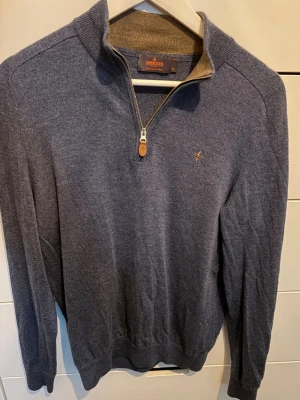 Blå stickad tröja med halvzip Morris - Snygg blå stickad tröja från Morris med halvzip och hög krage. Tröjan har diskret logobrodyr på bröstet och ribbade muddar vid ärmslut och nederkant. Perfekt för lager-på-lager och passar till jeans eller chinos