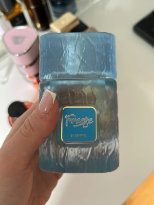 Freeze parfym från RiiFFS blå - Säljer en Freeze parfym från RiiFFS i en unik, frostad blå glasflaska med struktur och fyrkantig form. Flaskan har ett blått och guldigt emblem på framsidan och ett matchande blått lock. Perfekt för dig som gillar snygg design och fräscha dofter. Oanvänd. 
