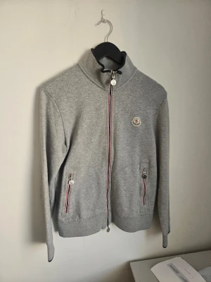 Grå zip hoodie från Moncler - Snygg grå hoodie från Moncler med dragkedja framtill och två fickor med randiga detaljer i rött, vitt och blått. Moncler-logga på bröstet och hög krage med svart kant. Tillverkad i mjukt sweatshirtmaterial, perfekt för en chill och stilren look.orginal M passar S bättre