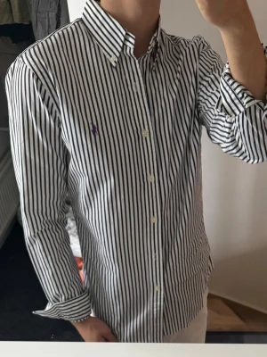 Randig skjorta från Ralph Lauren - Säljer en klassisk vit skjorta med svarta vertikala ränder från Ralph Lauren. Skjortan har lång ärm, knappar hela vägen och en lila broderad logga på bröstet. Tillverkad i bomull och har en snygg button-down krage. Perfekt för dig som gillar stilrena plagg.