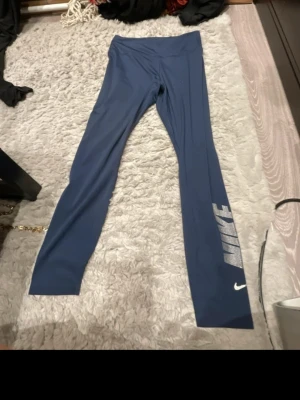 Blå Nike leggings med logga - Snygga blå leggings från Nike med stor vit logga längs ena benet. Tillverkade i ett stretchigt material av 79% polyester och 21% elastan som ger en skön passform. Perfekta för träning eller chill. Hög midja och långa ben.