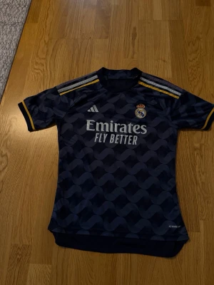 Real Madrid matchtröja Adidas blå - Snygg blå Real Madrid matchtröja från Adidas med gulddetaljer på axlarna och klubbmärke på bröstet. Tillverkad i lätt och ventilerande material, perfekt för fotbollsträning eller match. Tryck med 'Emirates Fly Better' på framsidan.