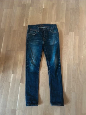 Mörkblå jeans från Nudie Jeans - Säljer ett par mörkblå jeans från Nudie Jeans med klassisk femficksmodell och smal passform. Jeansen har snygga slitningar och kontrastsömmar, samt bälteshällor och knappgylf. Perfekta för en avslappnad och trendig stil.