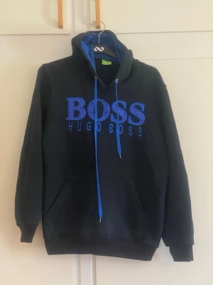 Svart hoodie från Hugo Boss - Säljer en svart hoodie från Hugo Boss med stor blå logga och text på bröstet. Hoodien har blå dragsnören, känguruficka framtill och en klassisk huva. Perfekt för dig som gillar streetwear och vill ha något bekvämt och stilrent.