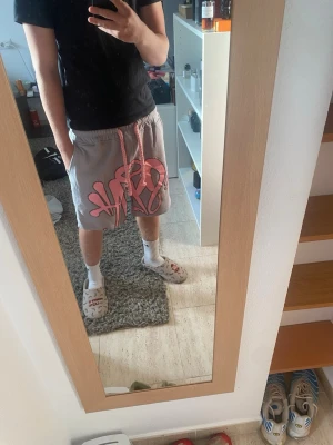 Synaworld shorts - Säljer ett par grå shorts med stort rosa graffiti-inspirerat tryck på ena benet och rosa snörning i midjan. Passar dig som är 160-180 Storlek S men är ganska stor för en S jag är 168 och den sitter så som ni ser på bilden på mig. Kom med frågor!