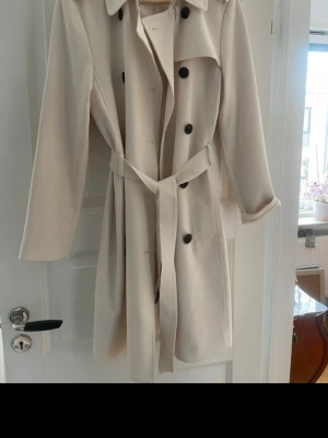 Beige trenchcoat från H&M - Stilren beige trenchcoat från H&M med klassiskt bälte i midjan och långa ärmar. Mjukt tyg som känns bekvämt.