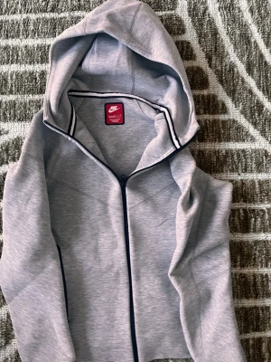 Grå zip hoodie från Nike - Snygg grå hoodie från Nike med dragkedja framtill och huva. Tillverkad i mjukt sweatshirtmaterial och har svarta detaljer längs dragkedjan och insidan av huvan. Perfekt för en sportig och avslappnad stil.
