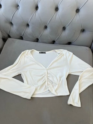 Vit croppad topp från Zara - Trendig vit croppad topp från Zara med lång ärm och v-ringning. Toppen har snygg knapprad framtill med rynkade detaljer som ger en cool twist. Perfekt att styla med höga jeans eller kjol för en fräsch look.