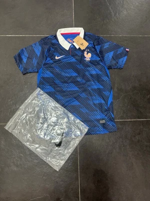 Blå Frankrike fotbollströja Nike - Säljer en blå fotbollströja från Nike med Frankrikes landslagsmärke på bröstet. Tröjan har ett mönster i olika blå nyanser, vit krage och korta ärmar. Perfekt för dig som älskar fotboll och vill stötta Frankrike på matchdagar.