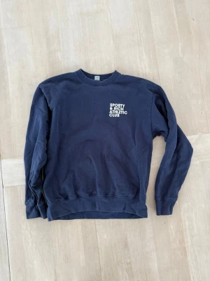 Mörkblå sweatshirt Sporty & Rich - Mörkblå sweatshirt från Sporty & Rich med vit text 'Sporty & Rich Athletic Club' tryckt på bröstet och ryggen. Klassisk rund halsringning, ribbade muddar vid ärmslut och nederkant. 