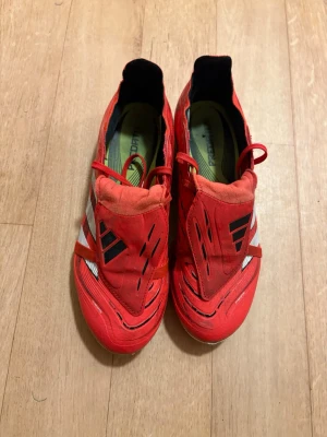 Adidas Predator röda fotbollsskor - Säljer ett par röda Adidas Predator fotbollsskor med vita och svarta detaljer. Skorna har klassisk låg profil, täckt snörning och greppvänliga dobbar på sulan. Tillverkade i syntetmaterial för bra bollkontroll och komfort på planen.