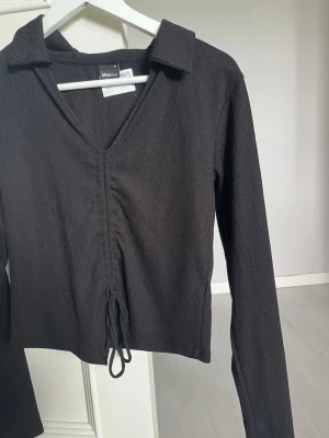 Svart långärmad topp från Dr. Denim - Snygg svart långärmad topp från Dr. Denim med krage och v-ringning. Toppen har snörning framtill som ger en cool detalj och croppad passform. Materialet är lätt ribbat och stretchigt, perfekt för att styla med jeans eller kjol.