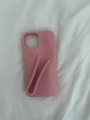 Rosa mobilskal från rhode med grepp till iphone 13 - Snyggt rosa mobilskal från rhode med unik greppfunktion på baksidan som gör det lättare att hålla i mobilen. Tillverkat i mjukt silikonmaterial som skyddar mot stötar och repor. Passar modeller med större kameralins. Perfekt för dig som vill ha både stil och funktion.