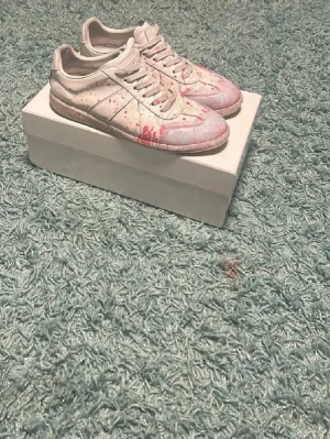 Maison Margiela sneakers med färgstänk - Unika sneakers från Maison Margiela i vitt läder med rosa och röda färgstänk. Klassisk låg siluett och diskreta sömmar. Snygg retrostil som sticker ut och passar perfekt till streetwear eller casual outfits. Skickar samma dag om inte man betalar på kvällen. Snabb affär bättre pris!