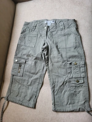 Khaki cargobyxor med fickor Wet Seal storlek 3US S - Säljer ett par khaki cargobyxor med flera praktiska fickor, dragkedjedetaljer och knappar. Byxorna har snörning vid bensluten och är tillverkade i ett mjukt bomullstyg. Perfekta för en avslappnad och street-inspirerad stil.