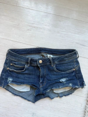 Mörkblå jeansshorts med hål - Säljer ett par mörkblå jeansshorts med fransiga kanter och slitna detaljer framtill. Strorlek 38 men sitter som 34/36. Klassisk femficksmodell med knappgylf och bälteshällor. Perfekta för sommaren och ger en avslappnad vibe.