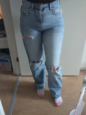 Ljusa slitna jeans - Säljer ett par ljusblå mom jeans med hög midja och raka ben. Jeansen har flera slitningar och hål på både fram- och baksida, vilket ger en trendig look. 
