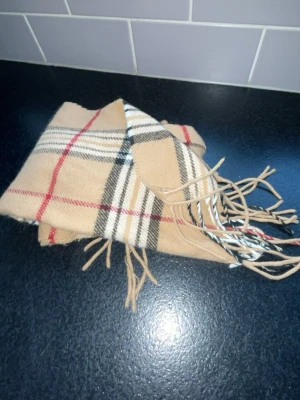Burberry scarf - Snygg beige halsduk med klassiskt rutmönster i svart, vitt och rött. Halsduken har fransar i ändarna och är perfekt att slänga över axlarna för en trendig look. Mjuk och skön, passar till de flesta outfits.