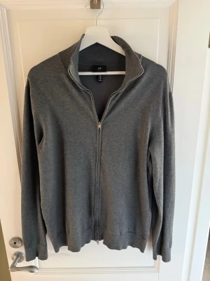 Stickad kofta  - Snygg grå fullzip från H&M. Tröjan har hög krage, hel dragkedja framtill och ribbade muddar vid ärmslut och nederkant. Perfekt för lager på lager och enkel att matcha med olika outfits. Mycket bra skick. Storlek M