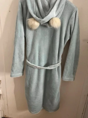 Ljusblå morgonrock med huva och öron  - Mysig ljusblå morgonrock i mjuk fleece med huva och två fluffiga vita bollar som dekoration. Rocken har knytskärp i midjan och två fickor framtill. Perfekt för lata morgnar eller kvällar hemma. Super skön och fin. Storlek 158/164 men passar i S också. Har bundle rabatt! 