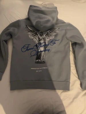 Grå hoodie från Hoodrich med ängelmotiv - Snygg grå hoodie från Hoodrich med stor ängelgrafik och texten 'From Nothing To Something' på ryggen. Framtill finns en mindre logga och ficka. Tillverkad i mjukt material och har en klassisk huva. Perfekt för dig som gillar streetwear och vill sticka ut.