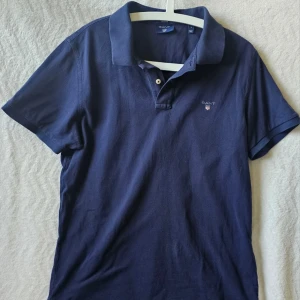 Marinblå pikétröja från Gant - Klassisk marinblå pikétröja från Gant med krage och knappar framtill. Tröjan har Gant-logga broderad på bröstet och är tillverkad i mjuk bomull. Perfekt till jeans eller shorts för en clean och stilren look.