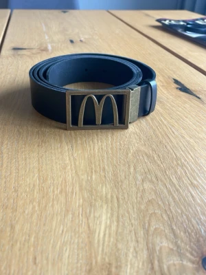Mc Donalds bälte  - Snyggt svart bälte med ett rektangulärt metallspänne som har en unik design. Bältet är stilrent och passar till många olika outfits. Perfekt accessoar för dig som vill ha något som sticker ut lite extra.