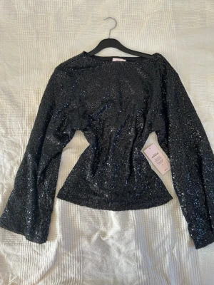 Svart glittrig topp med markerad midja och paljetter - Populär Snygg svart topp från Nelly täckt med glittriga paljetter. Modellen har långa ärmar och en figurnära passform som markerar midjan. Perfekt för fest och utgång! Är ganska stor i storleken så passar nog även xs/s💗