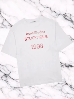 Vit Acne Studios t-shirt med tryck - Vit t-shirt från Acne Studios med rosa text 'Acne Studios STOCKHOLM 1996' på bröstet. Klassisk passform och rund halsringning, perfekt för en stilren och avslappnad look.