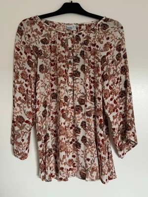 Blommig blus från Isolde - Luftig blus från Isolde i cremevitt tyg med blommigt mönster i bruna, röda och beige toner. Rund halsringning, knappar framtill och långa ärmar med lätt puff vid ärmslut. Perfekt för en avslappnad men stilren look.