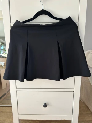 Svart kortkjol med veck - Säljer en svart kortkjol med snygga veck framtill. Kjolen har en lätt utsvängd form och är enkel att matcha till olika stilar. Perfekt för dig som gillar en klassisk men trendig look.