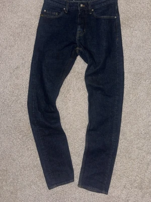 Mörkblå raka jeans från tiger of Sweden  Man - Snygga mörkblå jeans från tiger of Sweden  Man med klassisk femficksmodell och raka ben. Jeansen har en stilren look med bruna sömmar och patch med logga bak i midjan. Tillverkade i slitstark denim som passar perfekt till din vardagsstil.