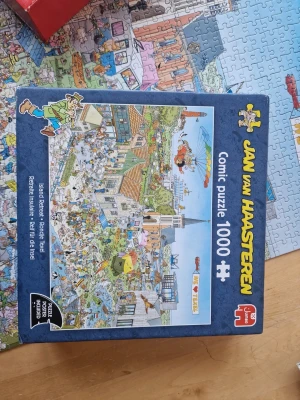 Jan van Haasteren 1000-bitar pussel - Komplett pussel med 1000 bitar från Jan van Haasteren. Motivet är tecknat i seriestil och föreställer en kaotisk och detaljrik stadsscen. Perfekt för dig som gillar utmanande pussel och humoristiska illustrationer.