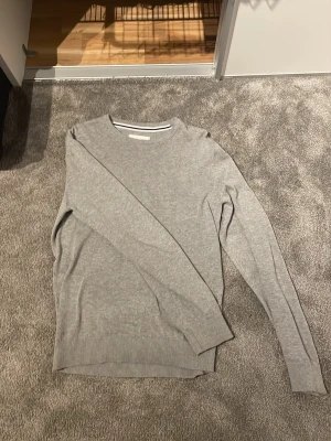 Ljusgrå stickad tröja - En stilren ljusgrå stickad tröja med rund halsringning och ribbade muddar vid ärmslut och nederkant. Perfekt basplagg som funkar till det mesta och är lätt att matcha. Materialet känns mjukt och smidigt.