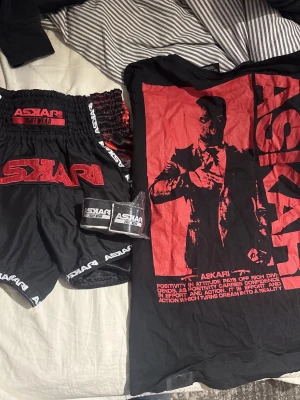 Svart och rött boxningsset från Askari - Snyggt boxningsset från Askari med svart t-shirt och shorts, båda med stora röda loggor och tryck. Setet inkluderar även svarta handlindor med vit logga. Perfekt för träning eller match inom kampsport. Thaiboxnings shorts med t shirt bindorna är 4,2M oöppnade, tröjan är M och byxorna är M skriv vid intresse mer bilder kan lösas. orginalpris på allt är 1500 mitt pris 800