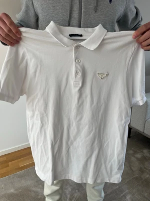 Vit pikétröja från Prada - Klassisk vit pikétröja från Prada med krage och knappar framtill. Tröjan har korta ärmar och en diskret logga på bröstet. Perfekt till jeans eller chinos för en clean och stilren look.