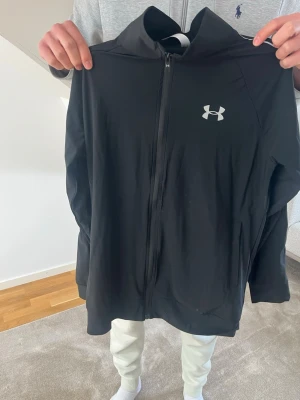 Svart träningsjacka Under Armour - Svart träningsjacka från Under Armour med hel dragkedja framtill och logga på bröstet. Tillverkad i lätt och funktionellt material som passar perfekt till träning eller uppvärmning. Jackan har hög krage och diskreta fickor på sidorna.