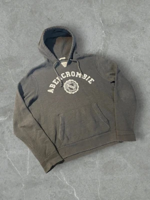 Mörkgrå hoodie från Abercrombie | L - Mörkgrå hoodie från Abercrombie med vit broderad logga och emblem på bröstet. Klassisk huva med dragsko och stor magficka framtill. Står XXL i men passar L