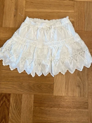 Vit kortkjol med spetsvolang - Supersöt vit kortkjol med bred resår i midjan och en volang. Kjolen har fina spetsdetaljer och broderade mönster. 