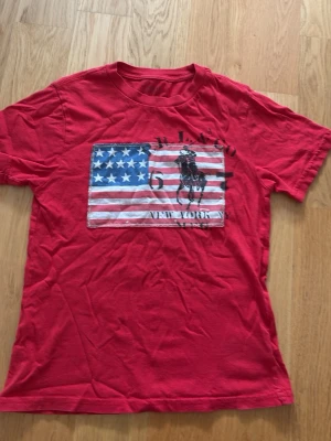 Röd t-shirt med USA-flagga från Polo Ralph Lauren - Snygg röd t-shirt från Polo Ralph Lauren med ett stort tryck av amerikanska flaggan. T-shirten har rund hals och korta ärmar, perfekt för en avslappnad streetstyle-look.