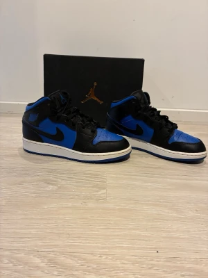 Air Jordan 1 Mid Svart/Blå - Snygga Air Jordan 1 Mid sneakers i svart och blått läder med klassisk Nike swoosh på sidan. Skorna har svart snörning, vit mellansula och blå yttersula. Perfekta för dig som vill ha en ikonisk och sportig look. 