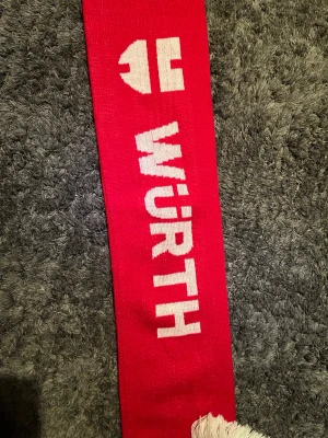 Röd FC Bayern München halsduk - Röd halsduk med FC Bayern München-logga och texten 'Würth' i vitt. Halsduken har vita fransar i ändarna och är perfekt för att visa ditt stöd för laget. Klassisk supporterdesign med tydliga klubbfärger.