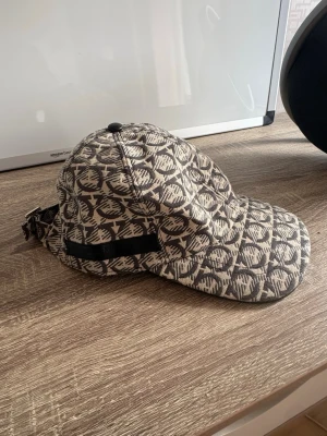 Ferragamo keps - Säljer en beige och svart keps med ett unikt grafiskt mönster över hela ytan. Kepsen har böjd skärm och justerbart band bak. Materialet ser ut att vara kraftig bomull eller canvas. Perfekt för dig som vill sticka ut med accessoarerna. Kepsen är onesize. 10/10 skick, använd bara ett fåtal gånger. Priset kan diskuteras!