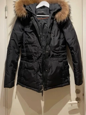 Svart dunjacka med pälskrage Hollies - Svart dunjacka från Hollies med stor luva och fluffig pälskant i brunt. Jackan har flera fickor framtill, dragkedja och knappar samt en figurnära passform. 